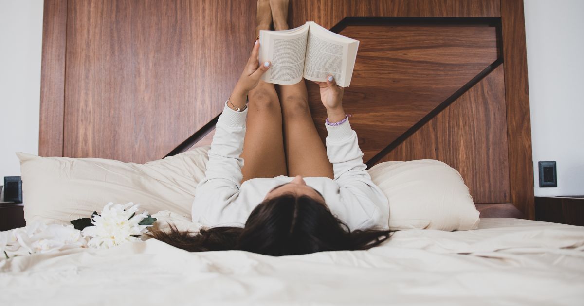 Young woman reading a book in bed with feet resting on the headboard 