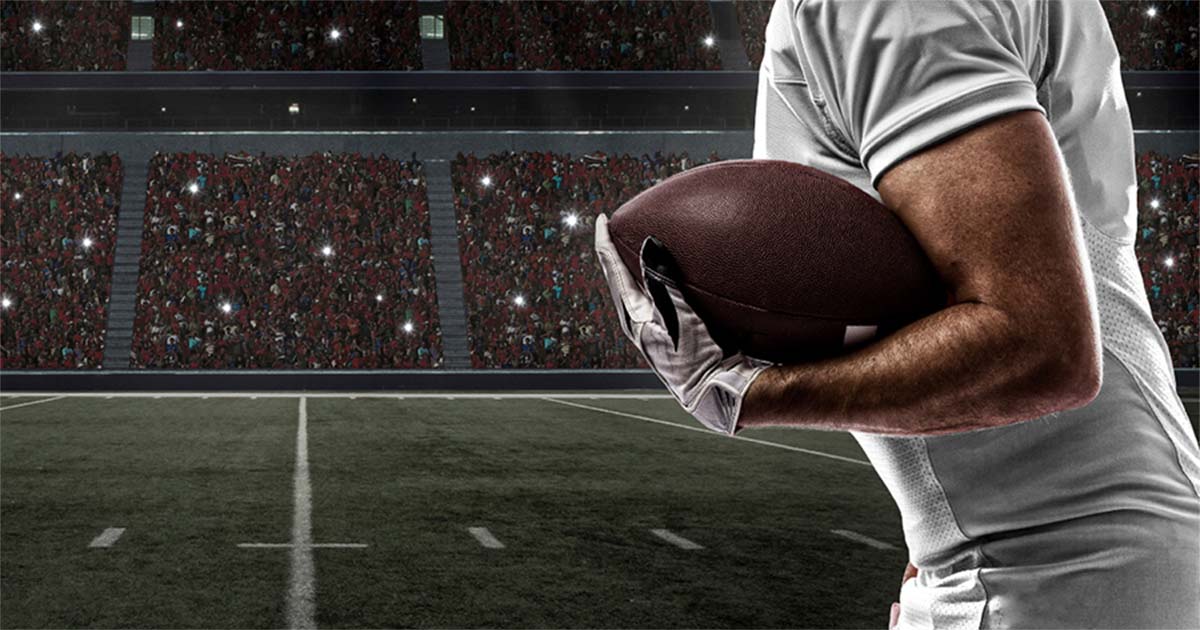 A photo of a football player cradling a ball. He is on the right, and there is a full stadium in the background.