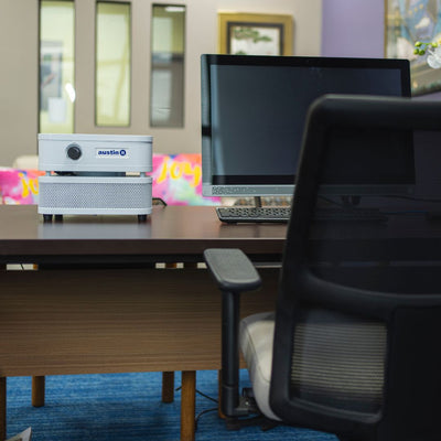 Austin air "it" on an office desk with computer setup, chair, and colorful decorations in the background